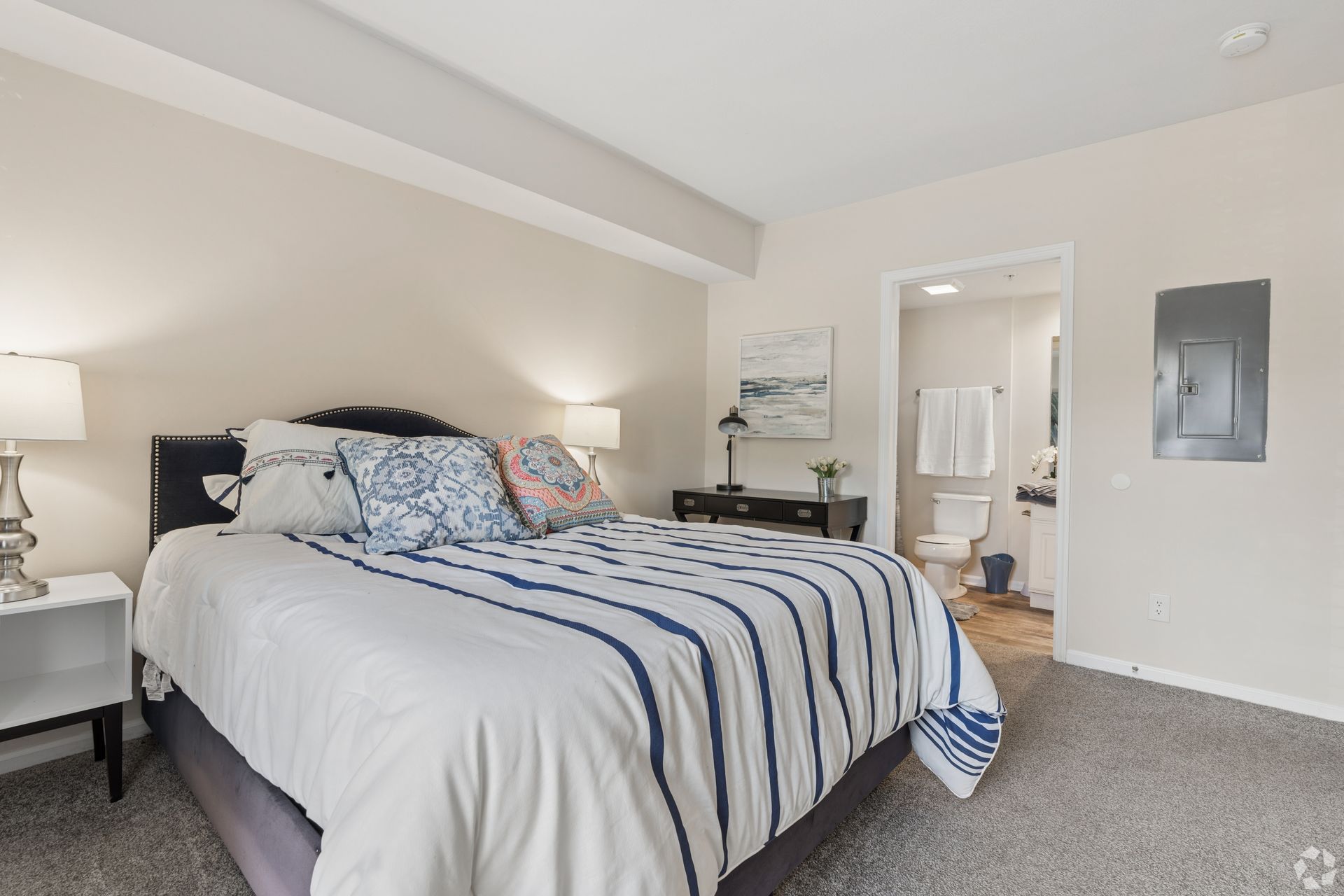 Bright bedroom with a neatly made bed, white bedding, blue striped blanket, and an open doorway to a bathroom at Quarry Apartments for Rent in Columbus, OH.