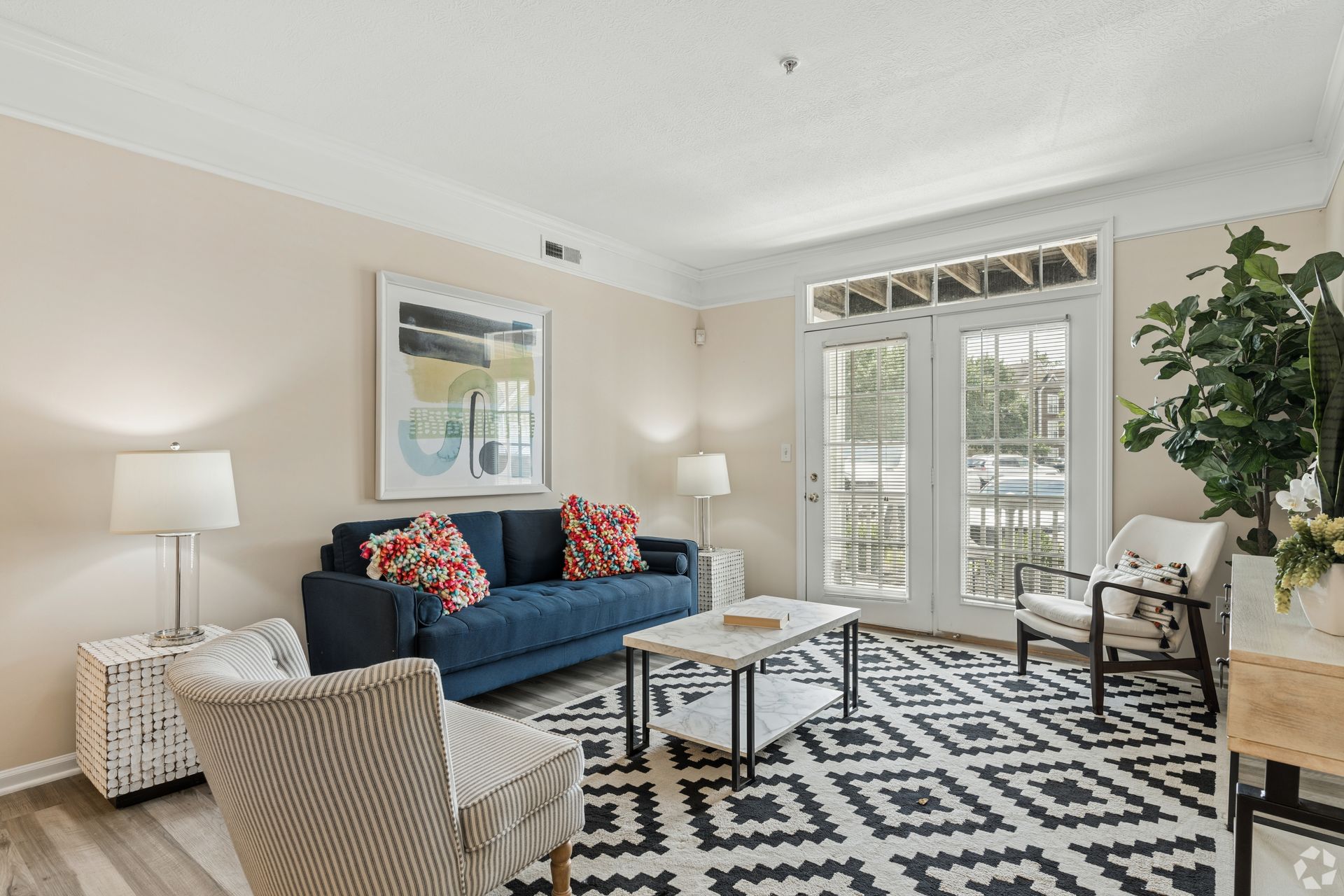 Bright living room with blue sofa, patterned rug, armchairs, lamp, and French doors with shutters at Quarry Apartments for Rent in Columbus, OH.