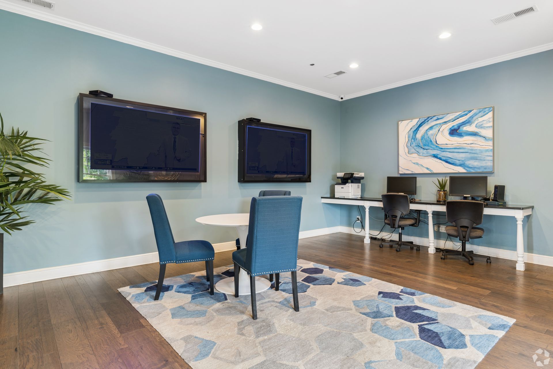 Modern home office with blue chairs, dual wall-mounted TVs, and a desk setup on hardwood floors.