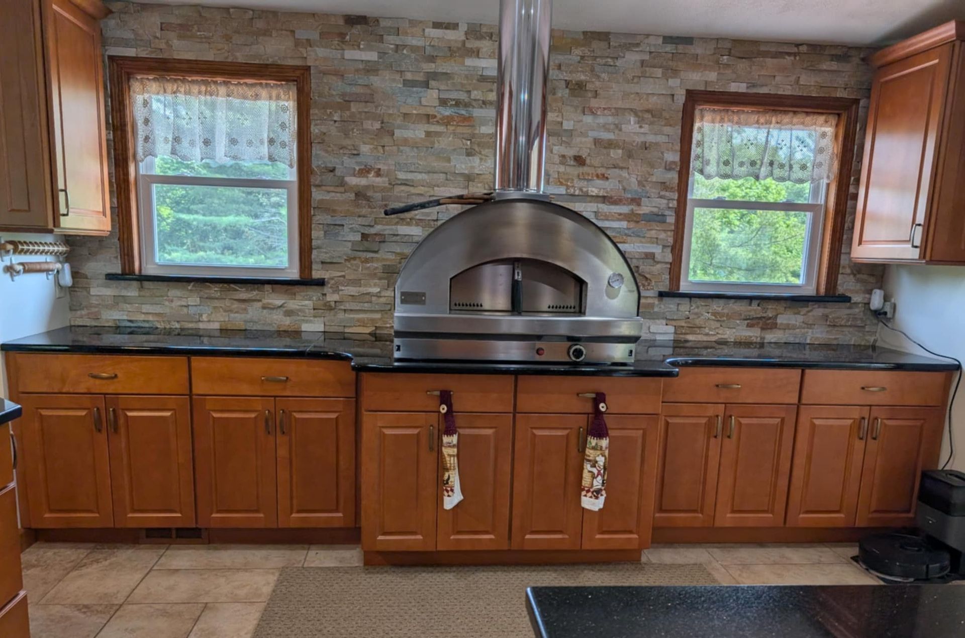 A kitchen with wood cabinets, black countertops, stone backsplash, and a large metal pizza oven with a vent hood.