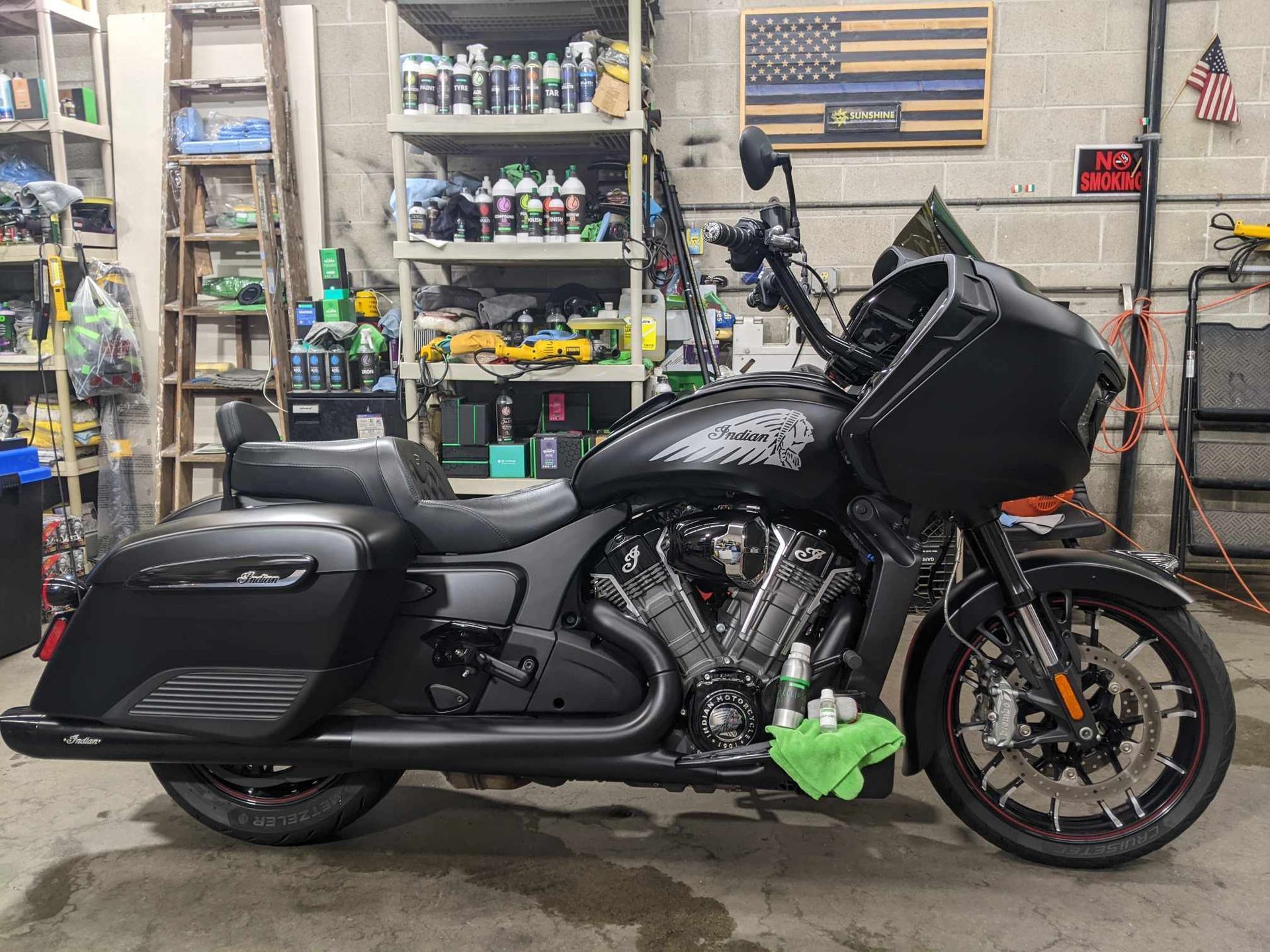 Matte black Indian Challenger motorcycle parked inside a garage workshop with shelves of supplies in the background.