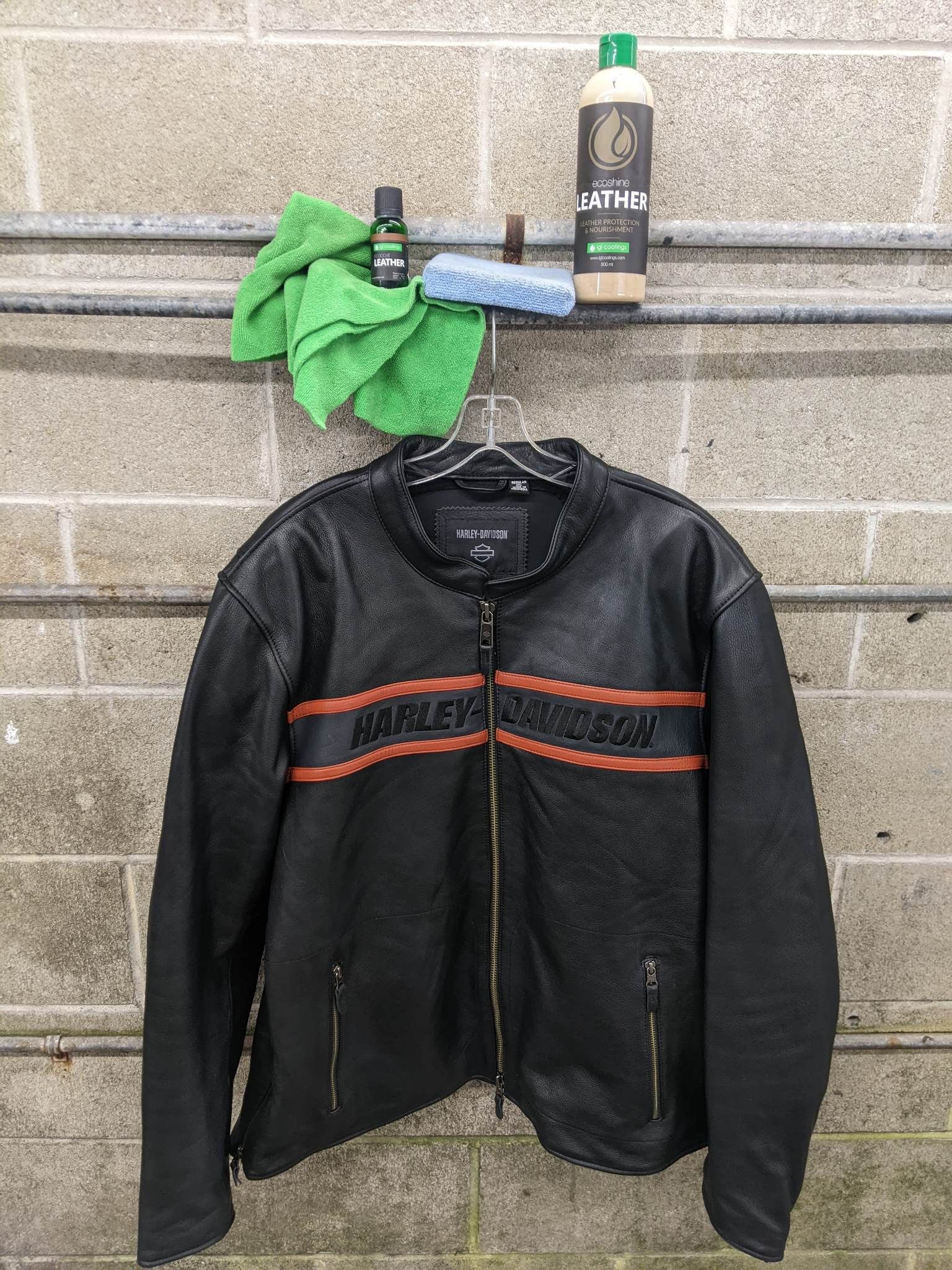 A black Harley-Davidson leather jacket hangs against a cinder block wall with leather cleaner, a sponge, and a cloth.
