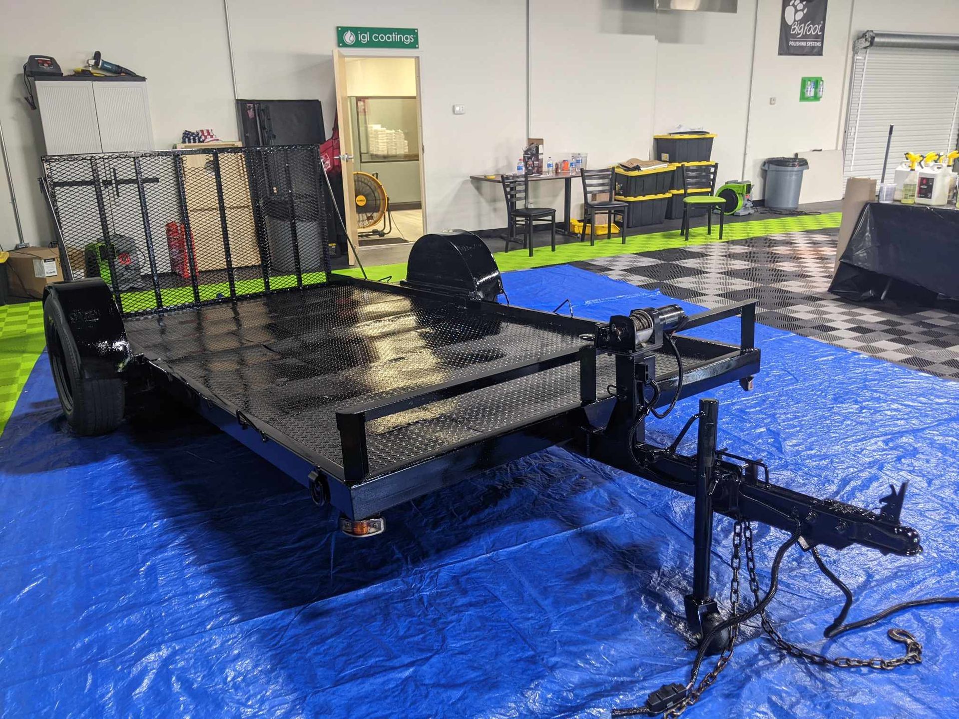 A black utility trailer with a diamond plate floor sits on a blue tarp inside a garage.