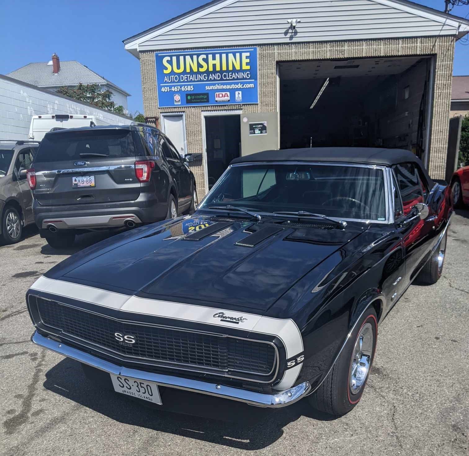 A black classic Chevrolet Camaro SS convertible parked in front of a Sunshine Auto Detailing shop on a sunny day.