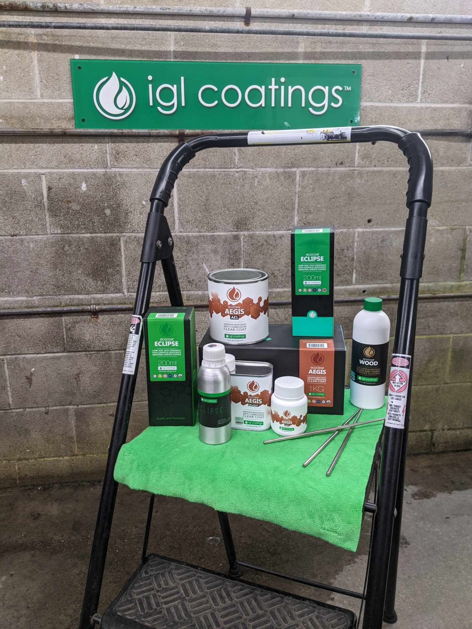 A collection of IGL Coatings products displayed on a step stool against a brick wall with a branded green sign.