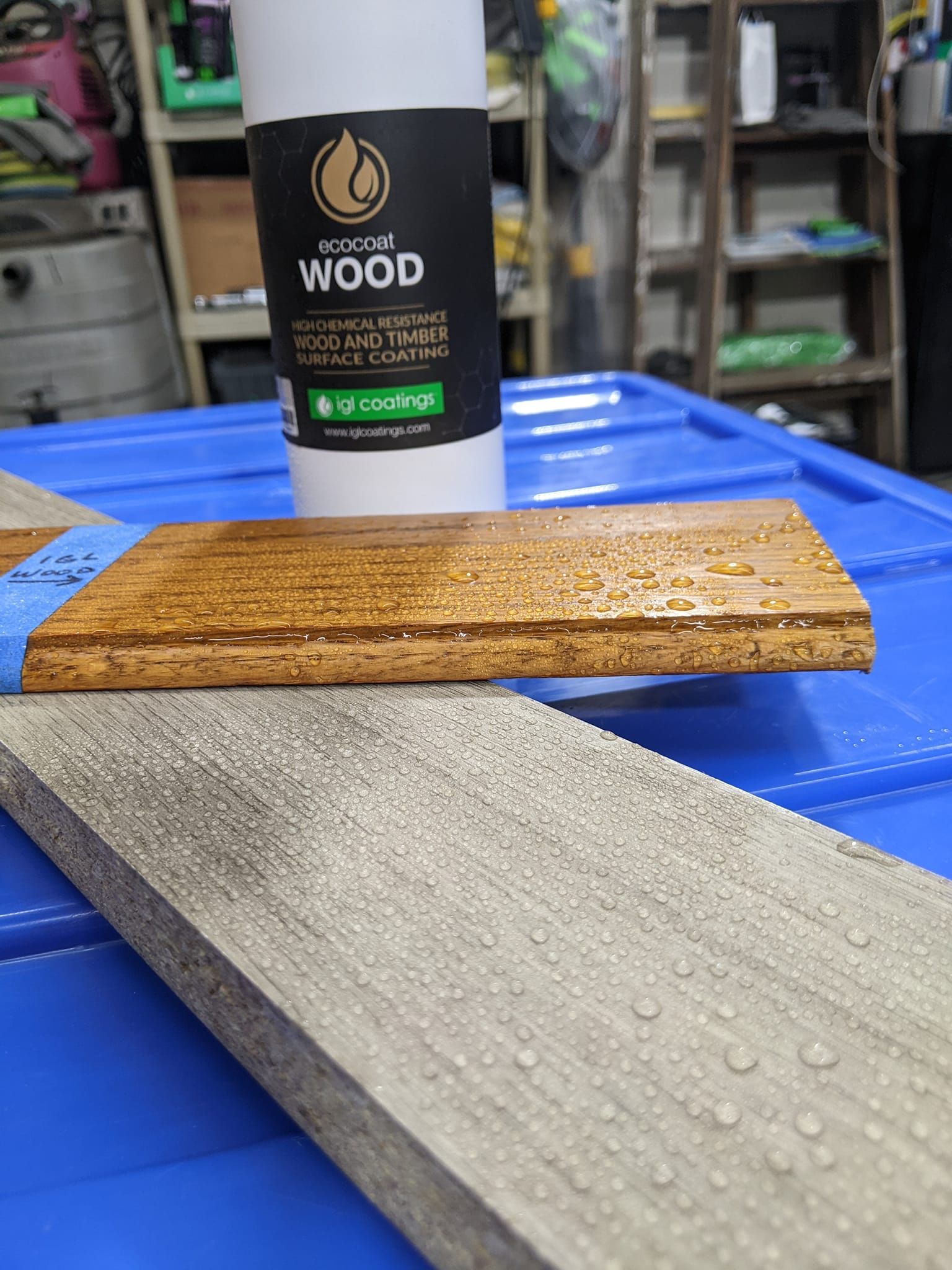 A bottle of wood protector sits behind a stained wood plank and an unfinished plank, both with water droplets on top.