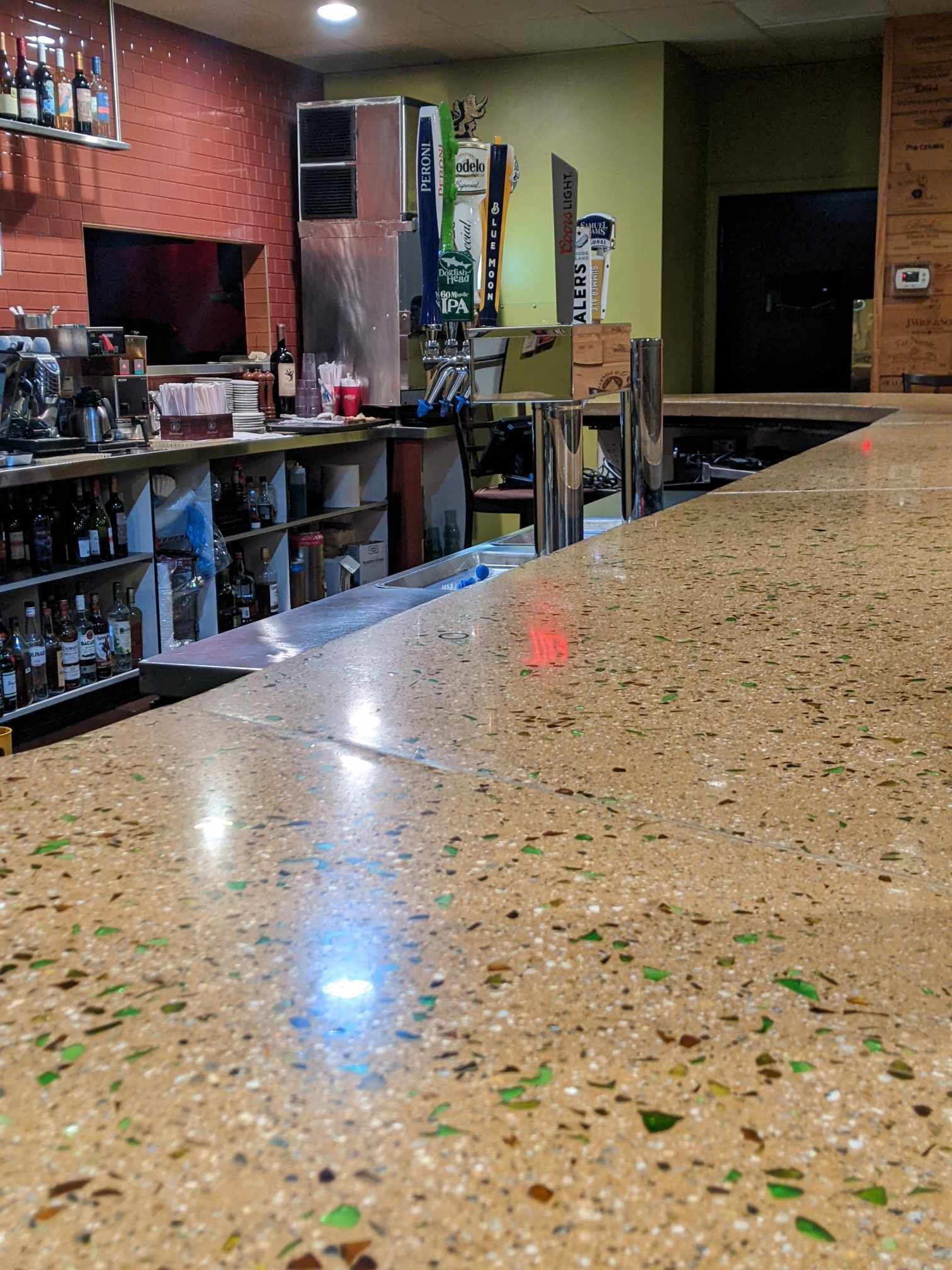 A close-up of a smooth, speckled bar countertop in a dimly lit bar, with a beverage station and bottles in the background.