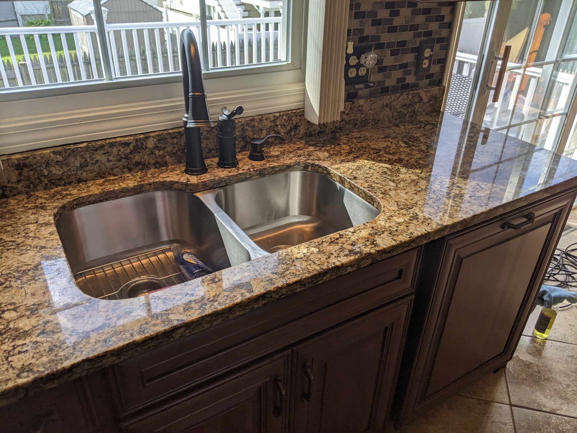 A stainless steel double-basin kitchen sink with a dark faucet set into a tan granite countertop above brown cabinets.