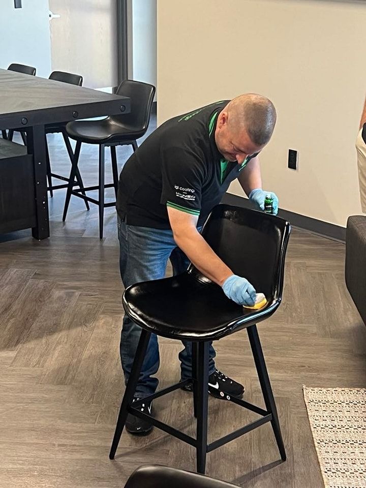 A person wearing blue gloves wipes down a black bar stool in an office setting.