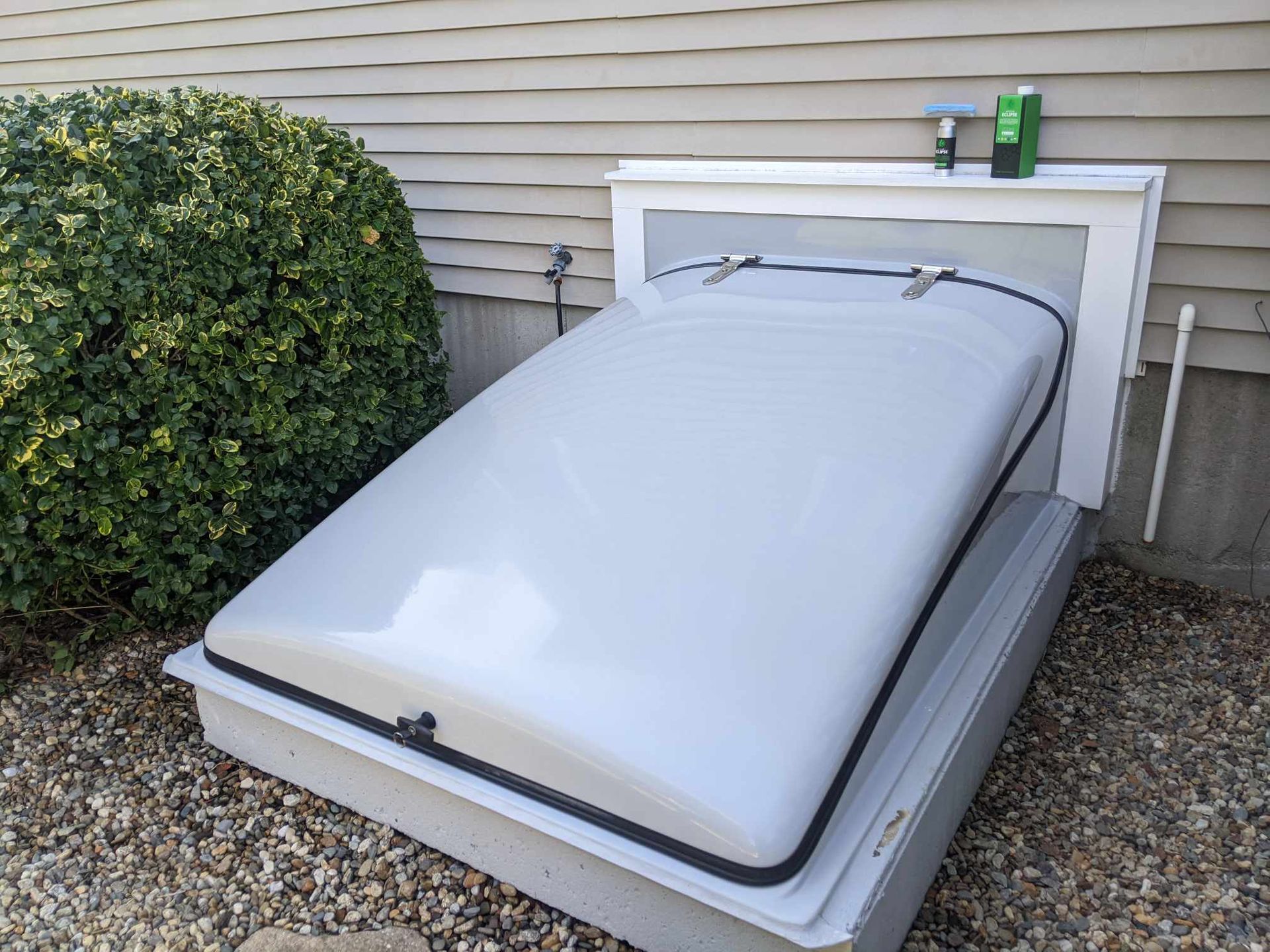A modern, grey basement bulkhead door installed on the exterior of a home next to a large bush and gravel ground.