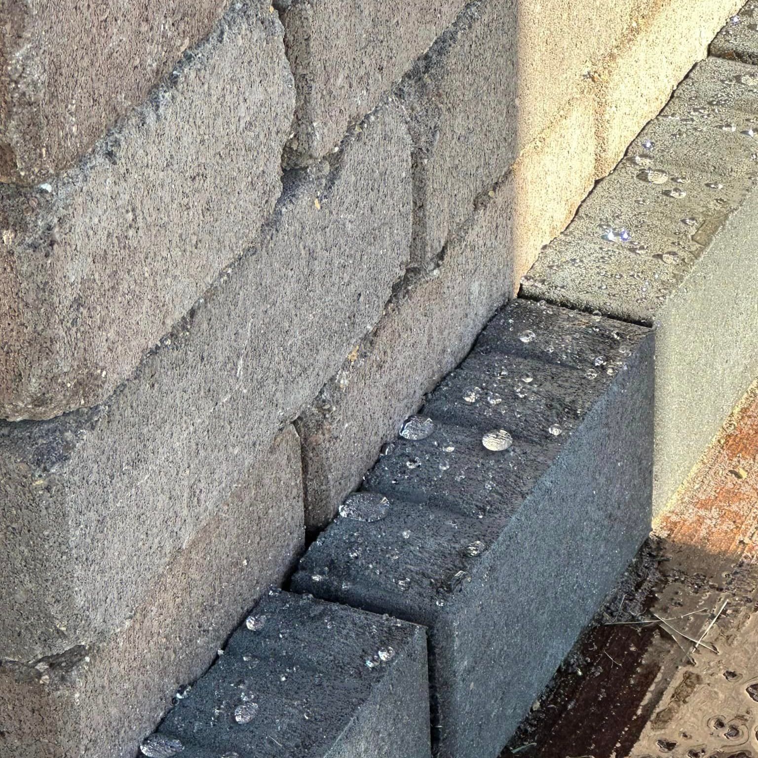 A close-up of textured grey masonry wall blocks meeting a row of darker rectangular pavers on the ground.