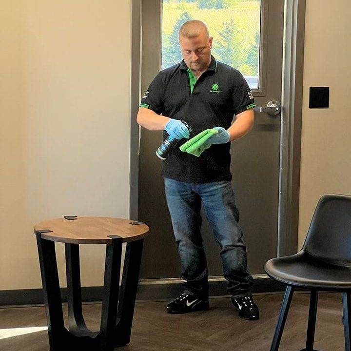 A person in a black polo and blue jeans sprays a cleaner onto a green cloth while standing near a modern side table.