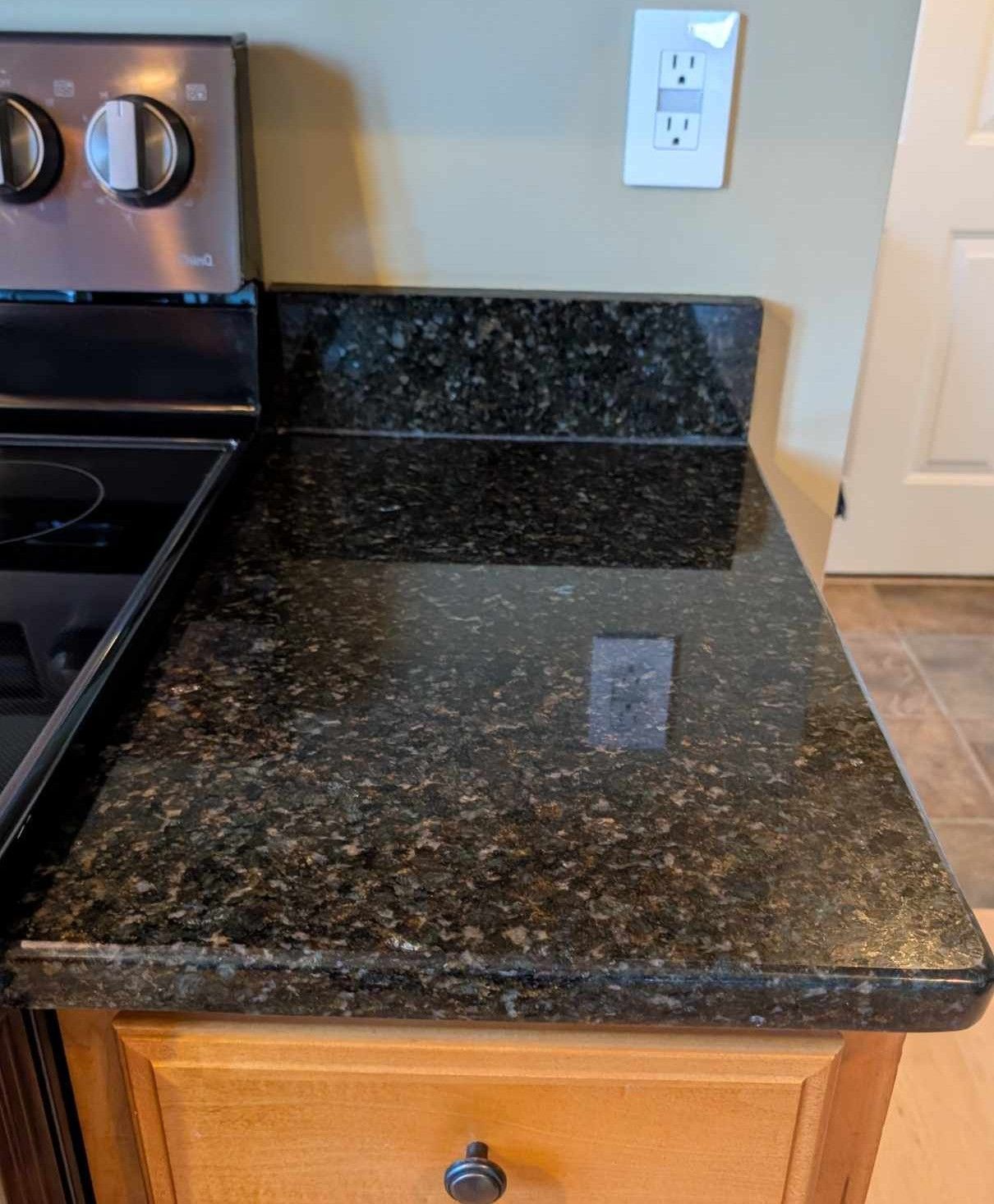 A kitchen counter with dark, speckled granite next to a stove, with a wooden cabinet and a wall outlet above it.