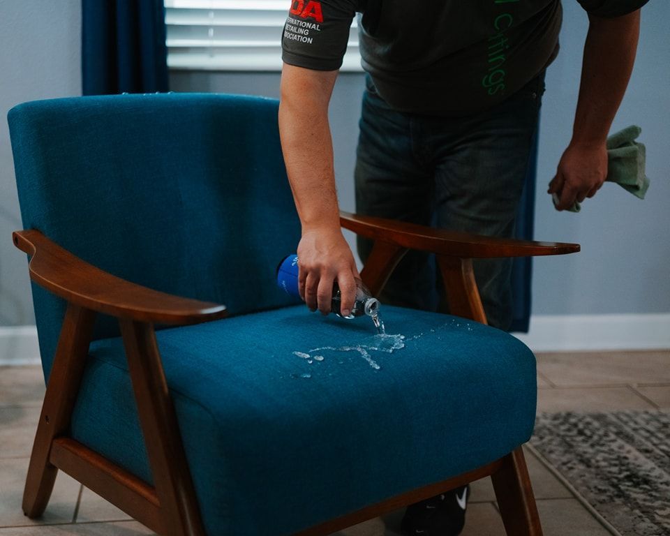 A person pouring liquid from a bottle onto the teal fabric of an armchair to demonstrate cleaning or spill resistance.