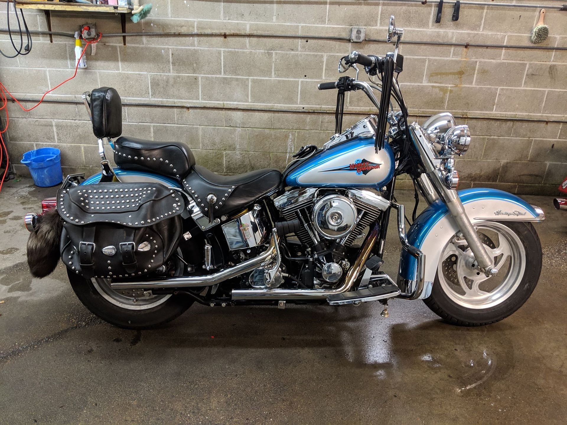 A blue and silver cruiser-style motorcycle with studded leather saddlebags and a backrest parked on a concrete floor.