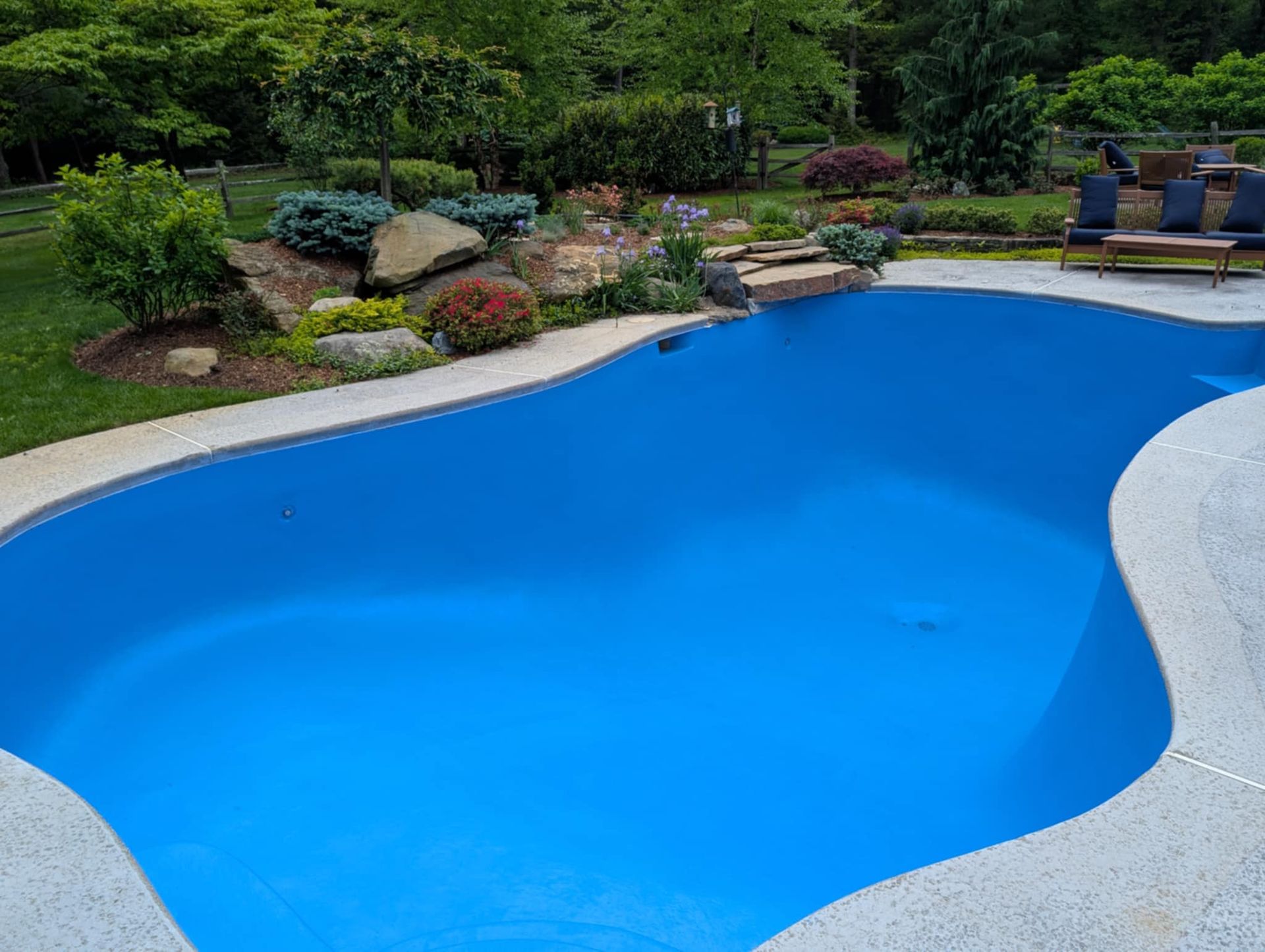 A curved, bright blue swimming pool with a textured stone coping, surrounded by a lush garden, rocks, and patio furniture.