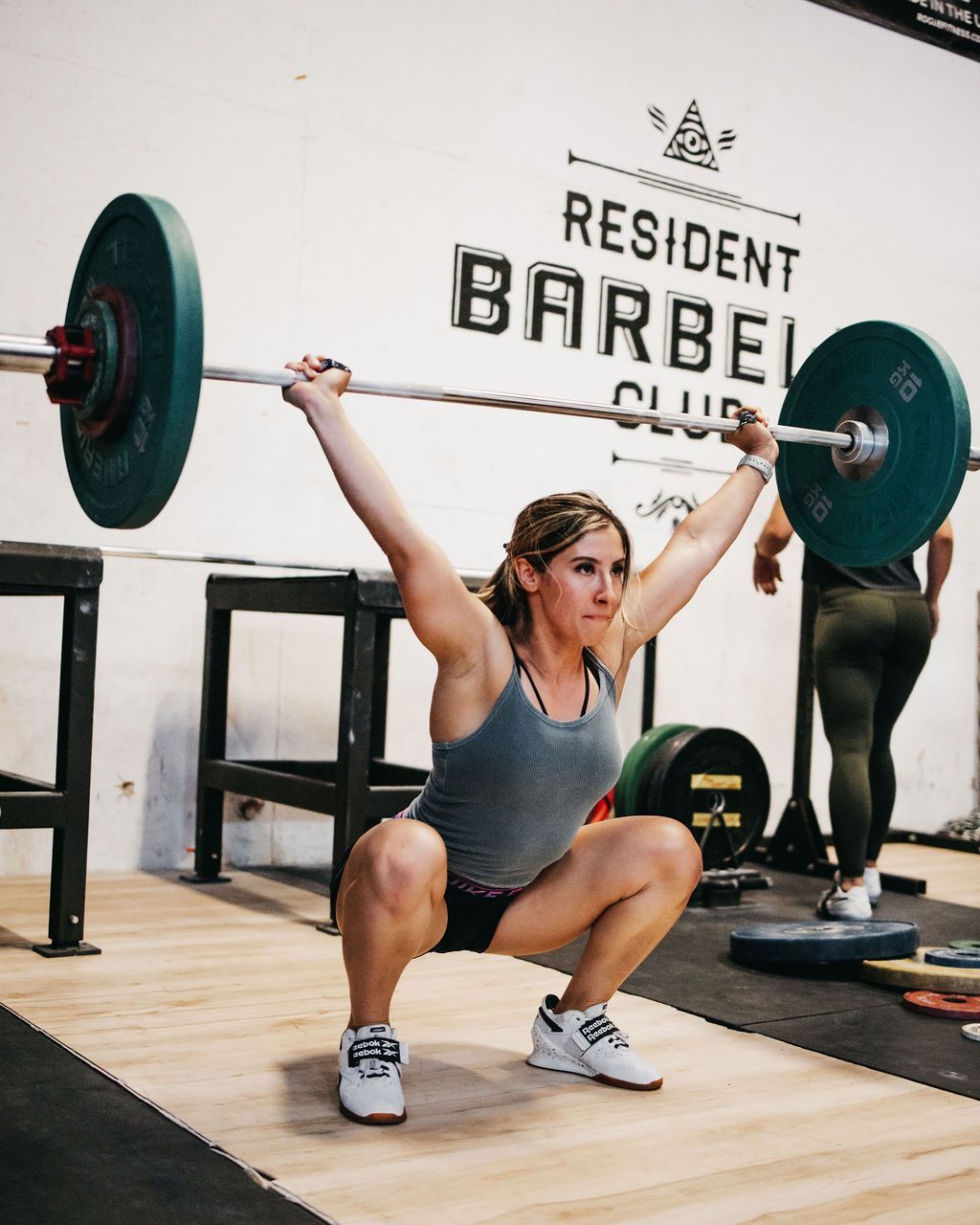 A woman squatting with a barbell over her head in front of a sign that says resident barbell club