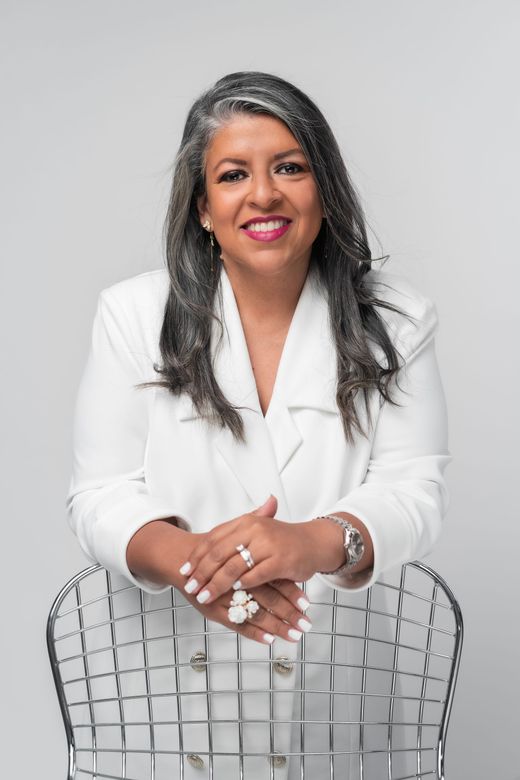 Woman in a white blazer smiles, seated with folded hands against a light gray background.