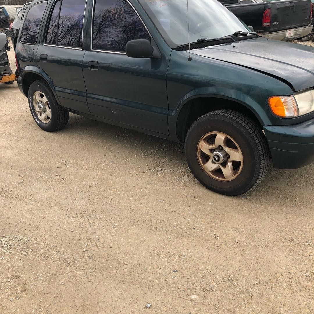 Green Kia SUV parked on a gravel surface. The car has tinted windows and a rusty wheel.