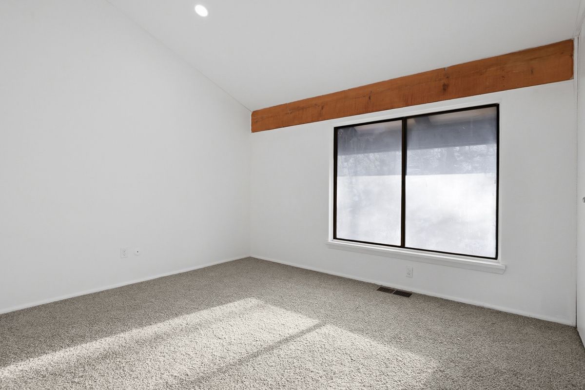 Empty room with white walls, carpet, and a window with a brown beam above it.