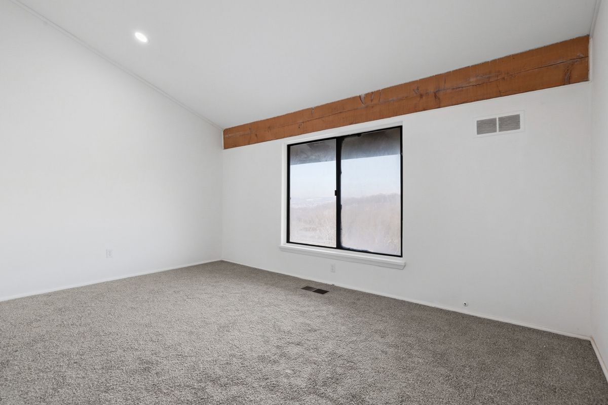 Empty room with white walls, angled ceiling, carpet, window, and a wooden beam.