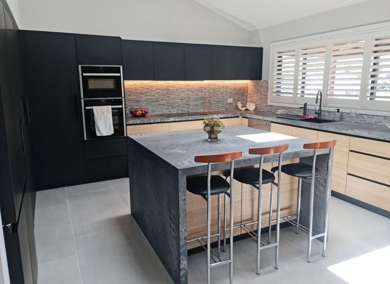 Modern kitchen with black cabinetry, stone countertop, island with bar stools, and shuttered window.