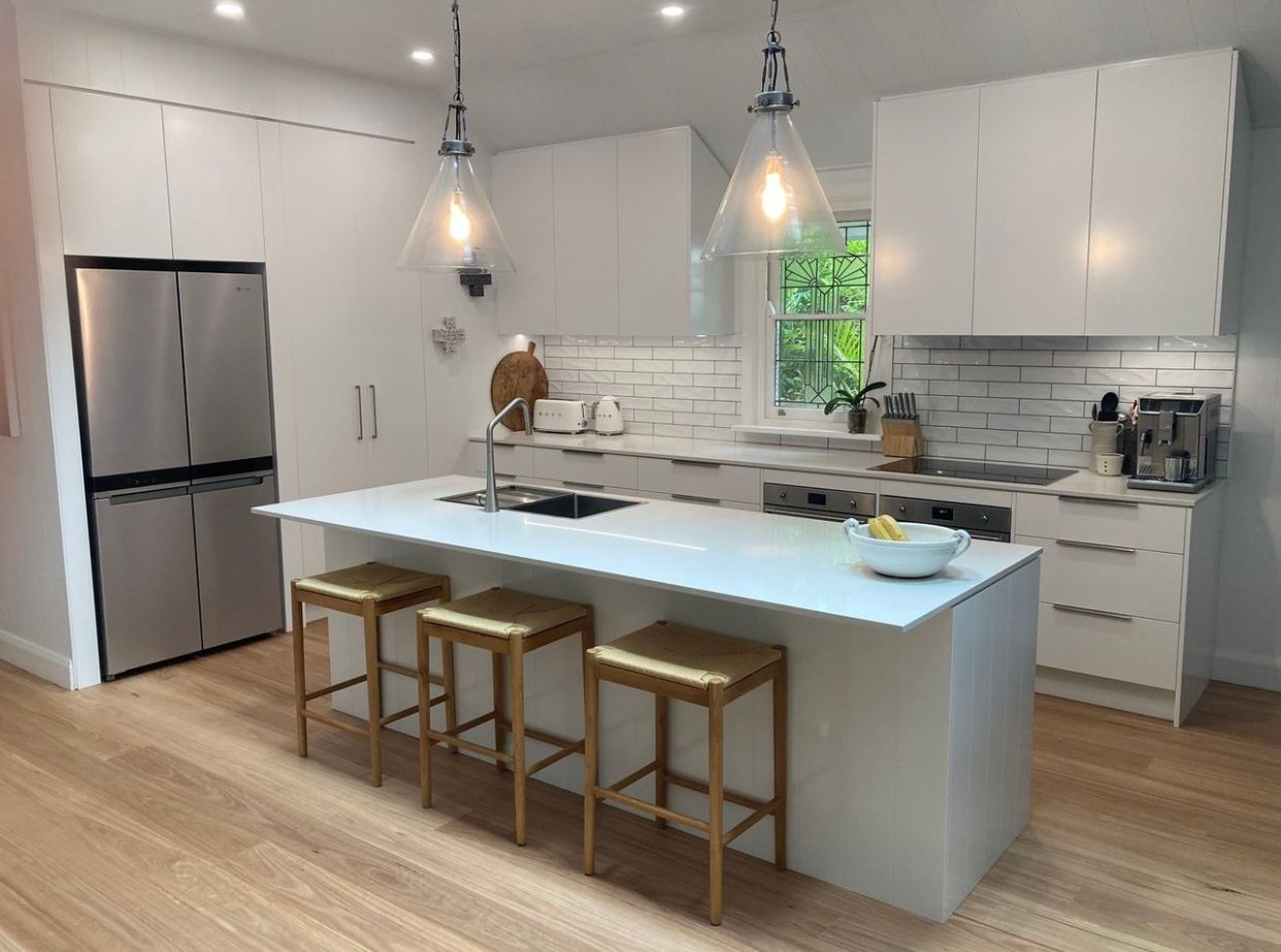Modern white kitchen with island, stainless steel fridge, and wooden stools.