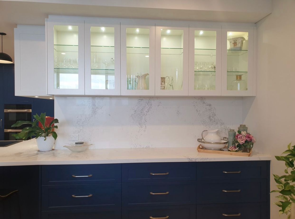 White glass-front cabinets above blue drawers in a kitchen, with a marble backsplash and countertop.