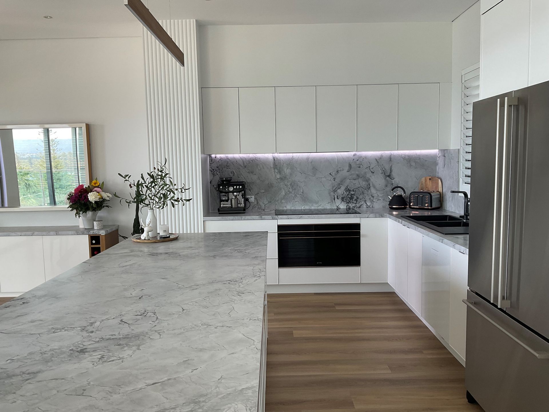 Modern white kitchen with granite countertops, stainless steel fridge, and wood flooring.