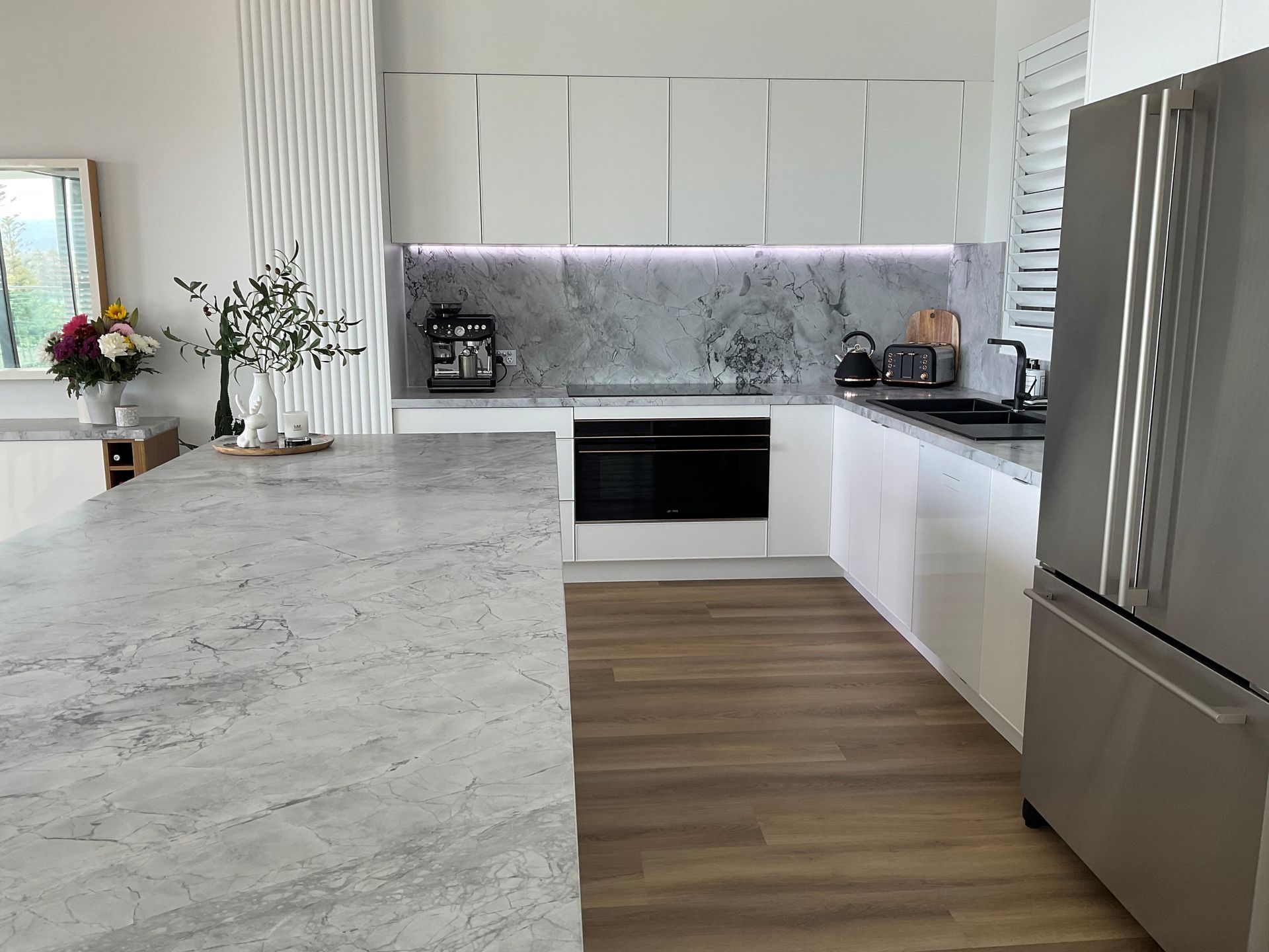 Modern white kitchen with marble countertops, stainless steel appliances, and wooden flooring.