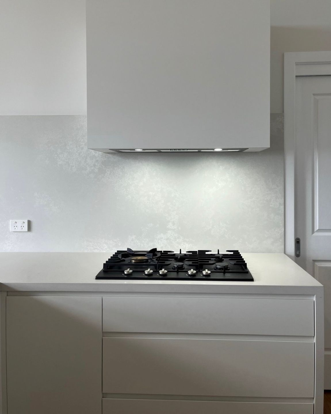 White kitchen with gas cooktop beneath a range hood; white countertop and cabinets; speckled backsplash.