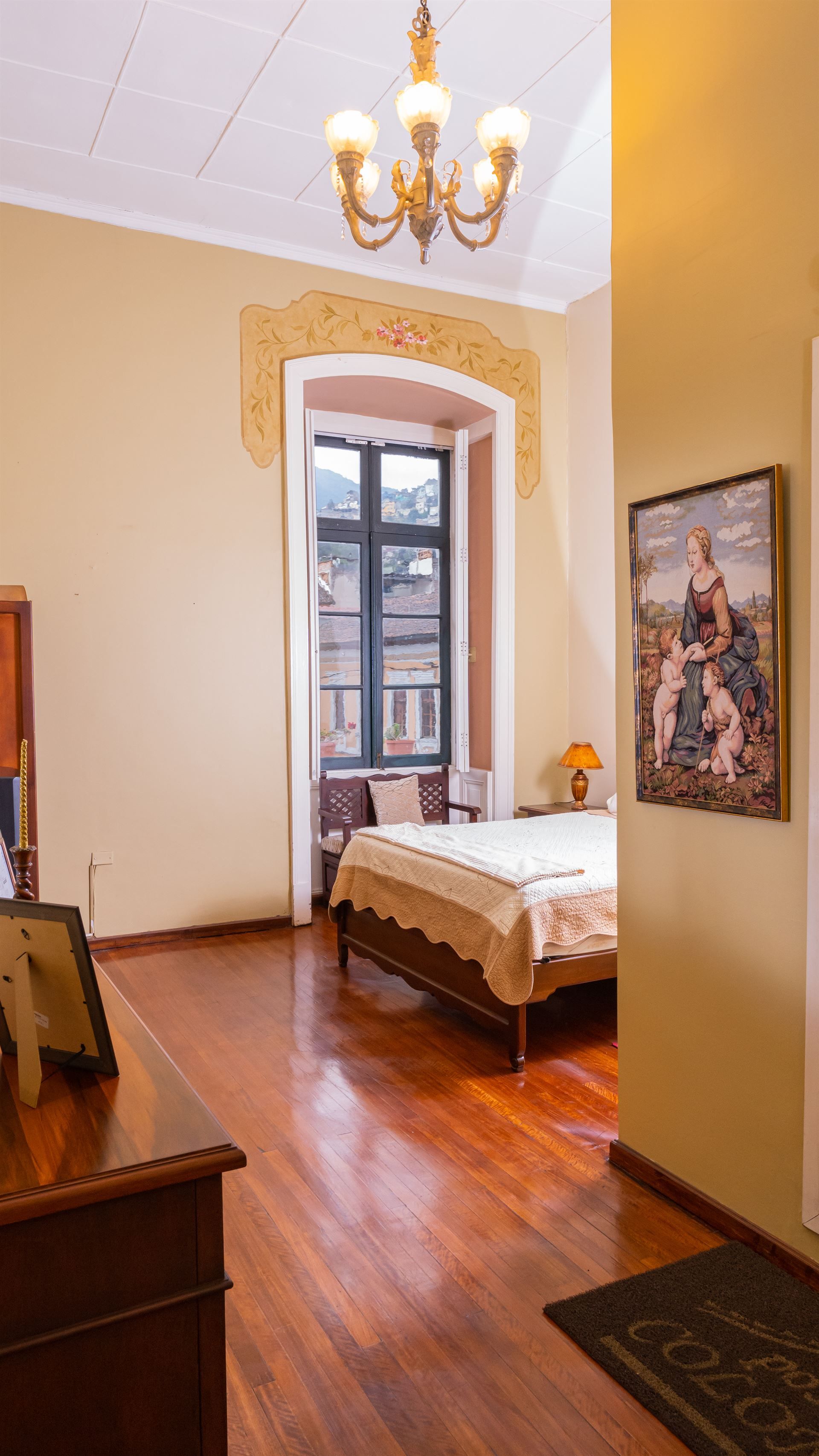 A bedroom with a bed , dresser , chandelier and painting on the wall.