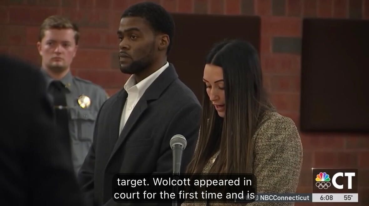 Courtroom scene with a man in a dark suit and a woman in a sweater standing before a judge.