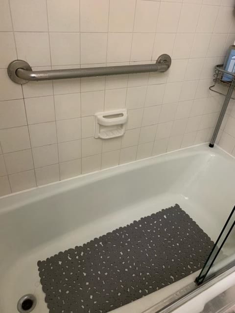 A bathtub with a gray mat on the floor and a handle on the wall.