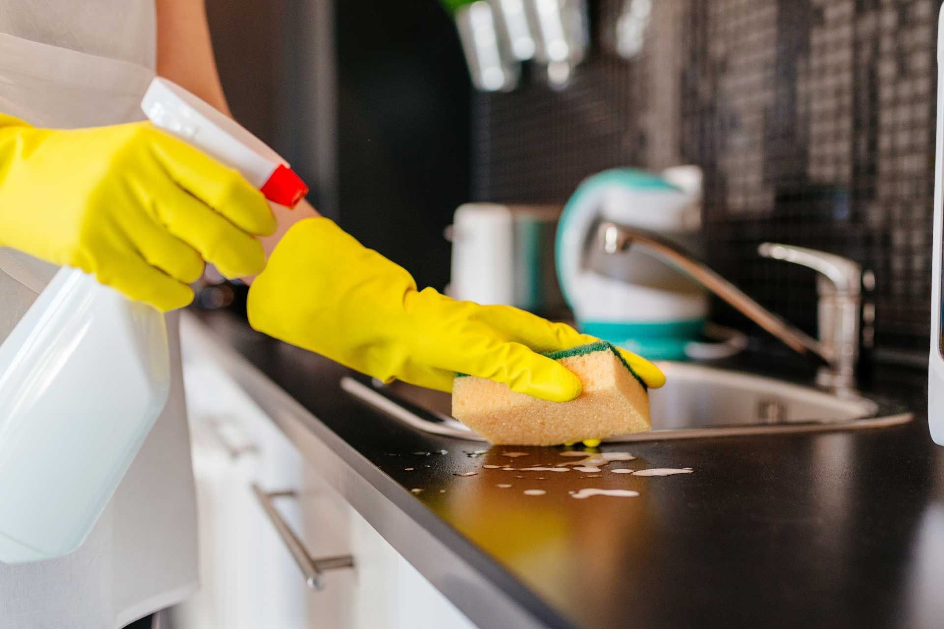 A person wearing yellow gloves is cleaning a kitchen counter with a sponge and spray bottle.