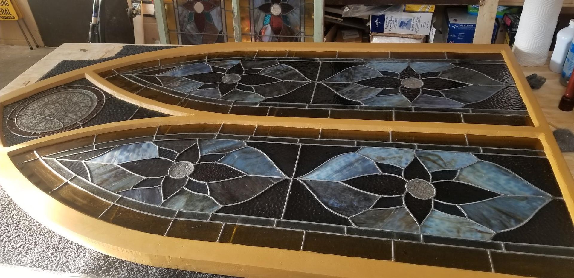 Two stained-glass window panels with blue, gray, and black floral patterns framed in wood, resting on a workshop table.