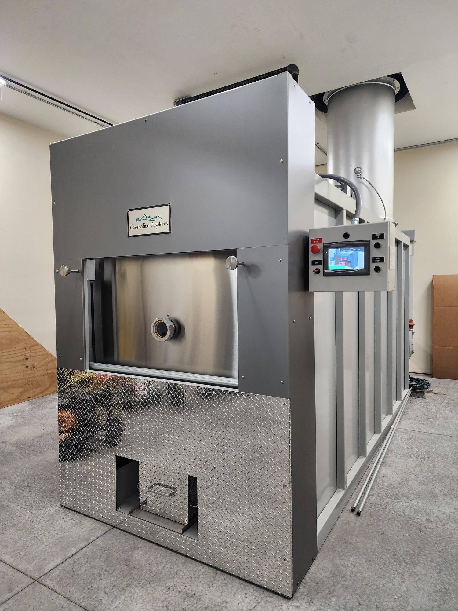 Industrial human crematory unit with a stainless steel door, diamond-plate base, and control screen, in an indoor space.