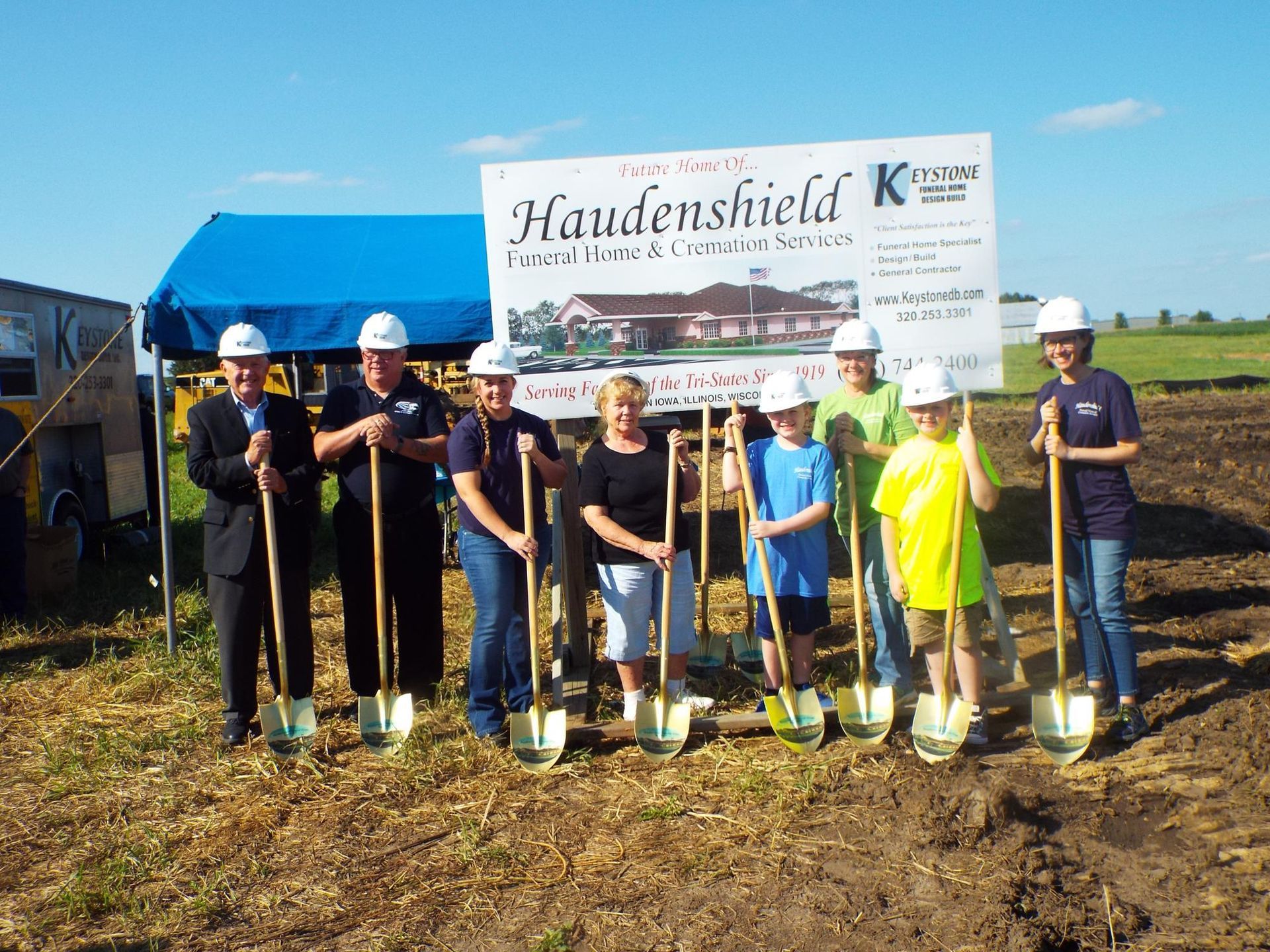 A group of people wearing hard hats hold shovels at a groundbreaking ceremony in front of a Haudenshield building sign.