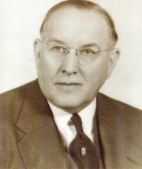 A portrait of a person in a suit, tie, and glasses, looking forward with a neutral expression.
