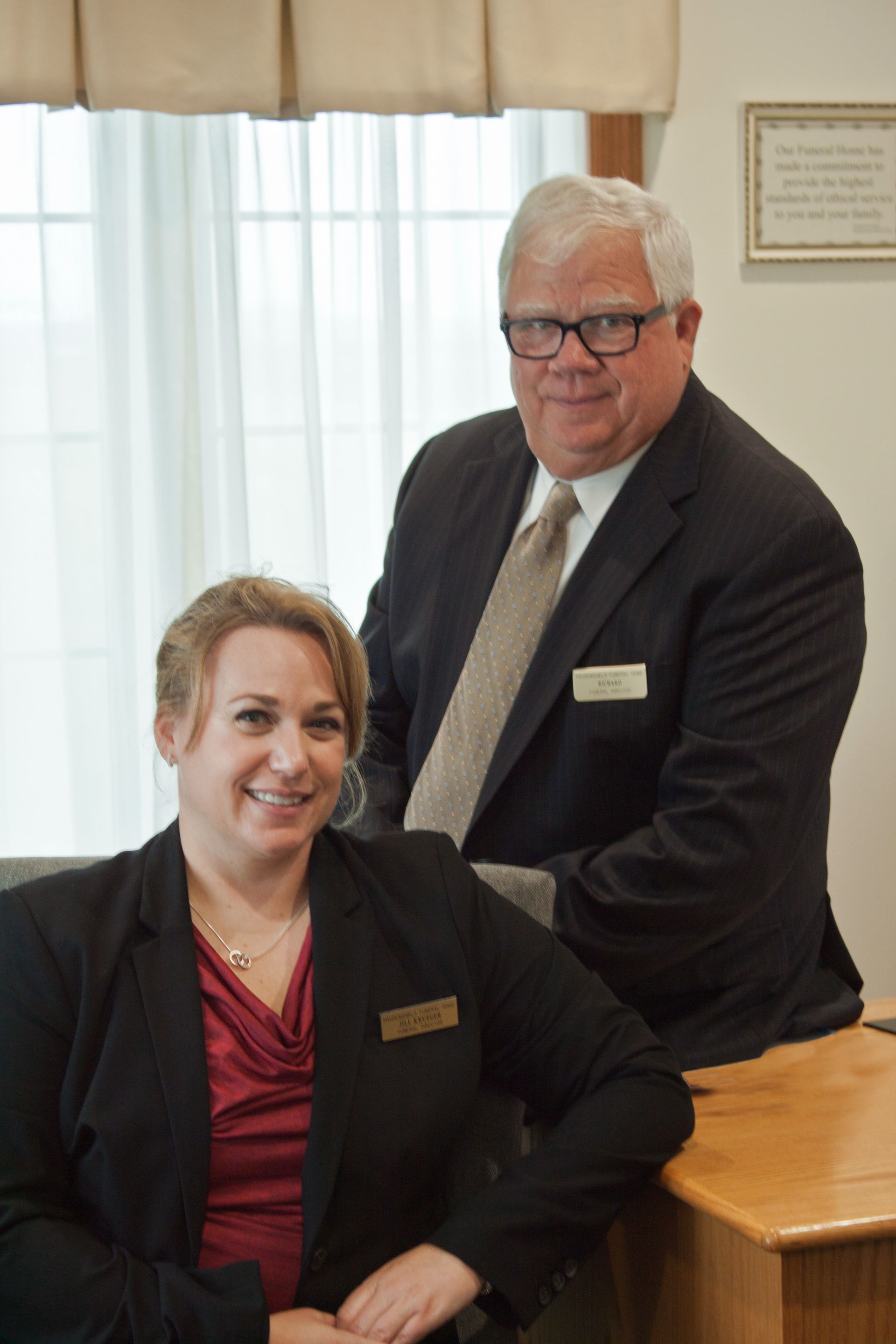 A woman in a black blazer and red top sits in front of a man in a suit and tie; both smile in a professional office.