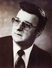 Portrait of a person in a suit and glasses, gazing slightly upwards with a neutral expression against a dark background.