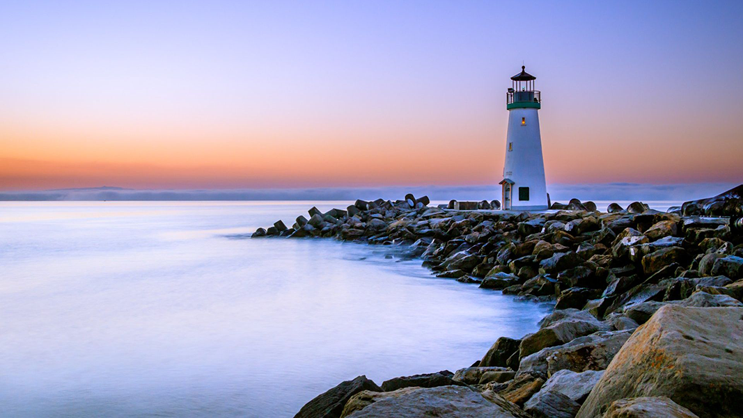 Lighthouse on rocky coast at sunset; white tower, blue water, orange and purple sky.