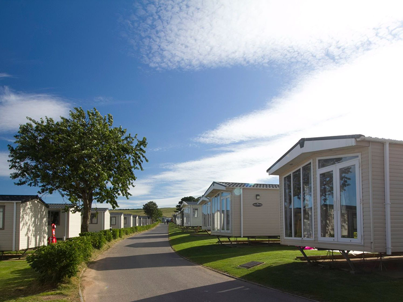 A row of beige mobile homes along a paved road under a blue sky with clouds.
