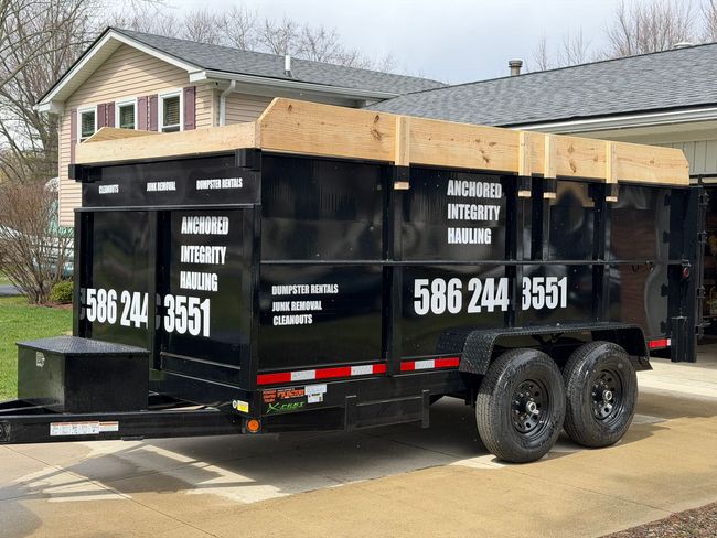 A black dump trailer parked on a concrete driveway in front of a house on a sunny day.
