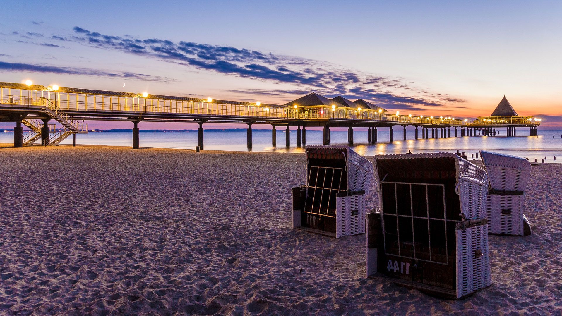Ein Strand mit Stühlen und einem Pier im Hintergrund bei Sonnenuntergang.