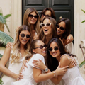 Six women in white outfits and sunglasses hug and smile outside a house.