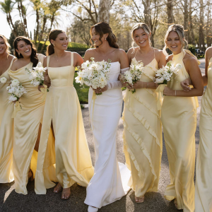 Bridesmaids in pale yellow dresses walk with a bride in a white gown outdoors, holding bouquets.