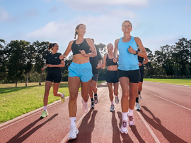 Group of runners jogging on a track outdoors in bright daylight, with trees and grass in the background.