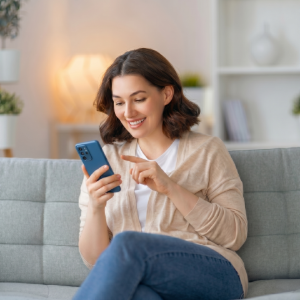 Woman smiling on a gray couch, using a blue smartphone in a bright living room.