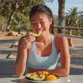 Woman in a light blue top smiling while eating melon at an outdoor table with fruit bowl